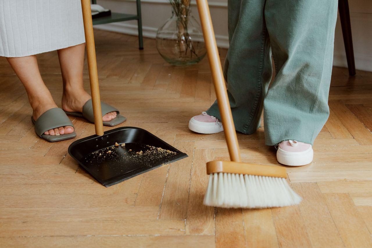 Close-up of people sweeping a wooden floor with a broom and dustpan indoors.