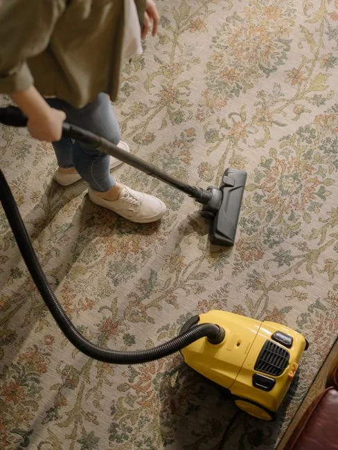 A woman vacuuming the floor while a dog looks on attentively. Indoor home setting.